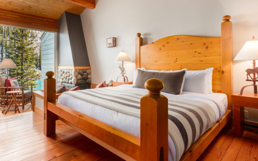 Cozy cabin interior at Moraine Lake with wooden beams, a double bed dressed in earthy-toned linens, a small seating area with armchairs, and large windows showcasing stunning views of the surrounding forest and mountains.