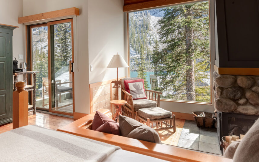 A cozy room at Moraine Lake Lodge featuring a bed with earthy linens, an armchair, and a dresser. The room is complemented by a stone fireplace and offers a scenic view of the surrounding nature.