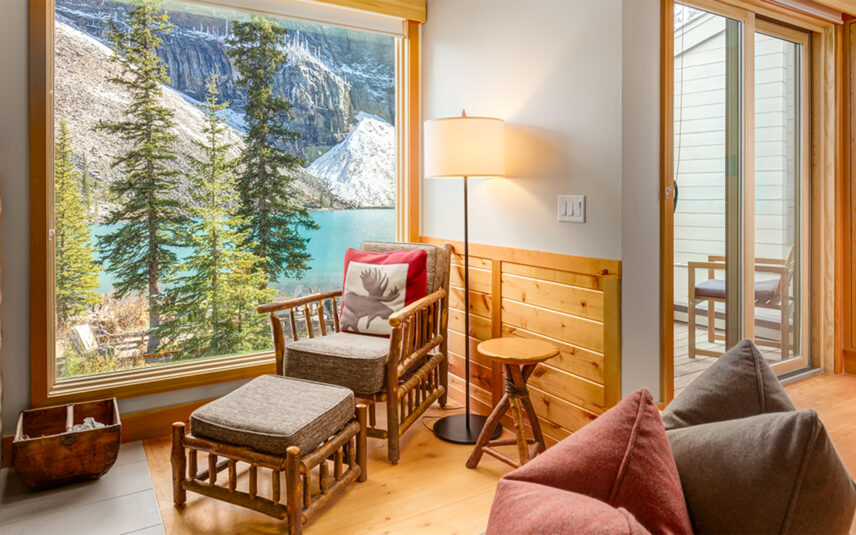 A lone armchair placed in front of a window with a stunning view of Moraine Lake, surrounded by towering mountains and lush trees, creating a serene and tranquil atmosphere.