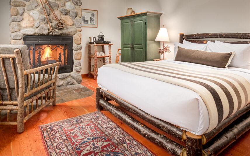 Inside a Moraine Lake room, a double bed with earthy linens sits next to a chest of drawers. A cozy seating area with armchairs and a warm fireplace add to the inviting atmosphere, perfect for enjoying a cup of coffee.