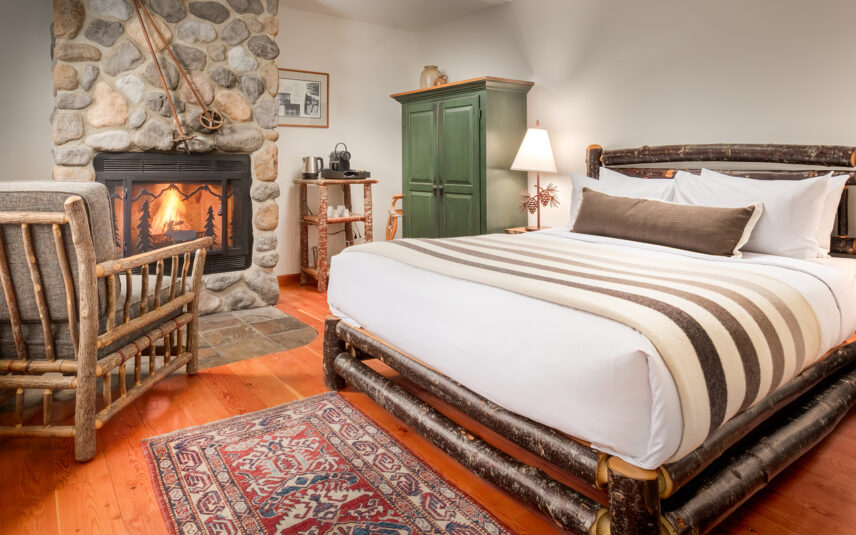 Inside a Moraine Lake room, a double bed with earthy linens sits next to a chest of drawers. A cozy seating area with armchairs and a warm fireplace add to the inviting atmosphere, perfect for enjoying a cup of coffee.