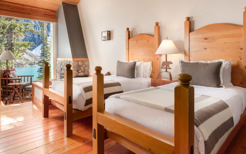Rustic cabin interior at Moraine Lake featuring two twin beds with earthy-toned linens, a small seating area with armchairs, and large windows showcasing scenic views of the forest and mountains.