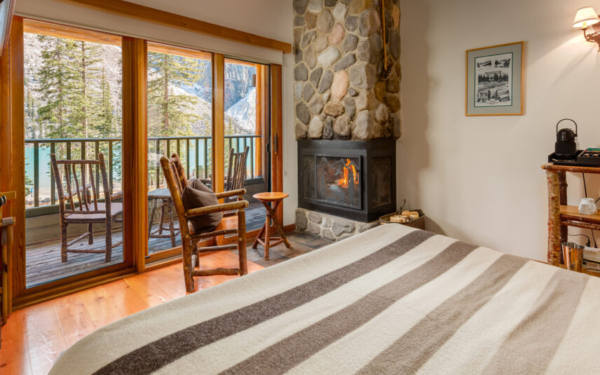A warmly lit lodge-style bedroom with wooden interiors and large windows looking out to a pine forest. The room features a modern bed with clean white linens and a striped throw, a stone fireplace with a cozy fire burning, and rustic wooden furniture. Natural light highlights the earthy tones of the hardwood floor and ceiling beams, creating a serene, mountain-retreat atmosphere.