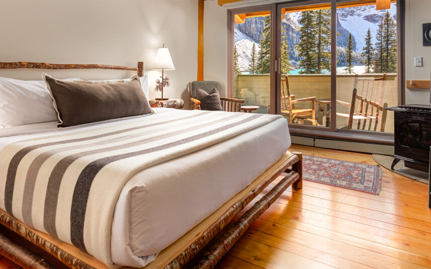 A cozy lodge-style bedroom featuring a king-sized bed with crisp white linens and a dark accent pillow. The room has warm wood accents throughout, including a wooden bed frame, ceiling beams, and flooring. A stone fireplace glows warmly beside the bed, and large windows let in natural light with views of the surrounding forest. A traditional patterned rug adds a touch of rustic charm to the space.