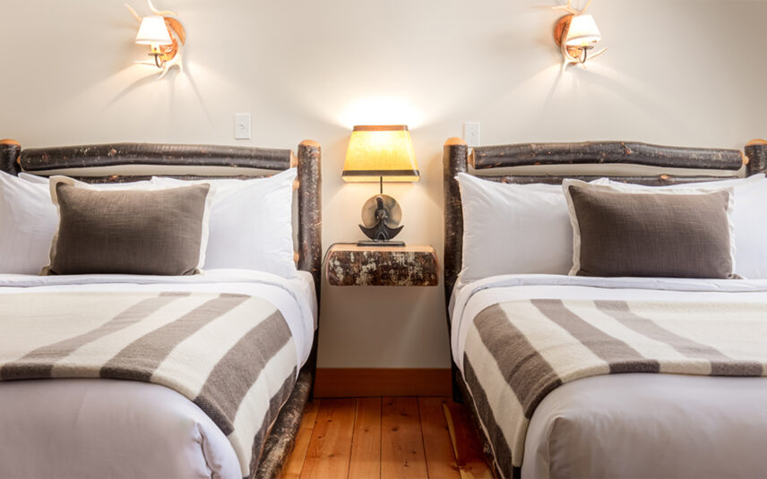 two twin beds lie side by side. Their frames are made of rugged wood, and they each have access to a rustic table with a warm lamp atop it.
