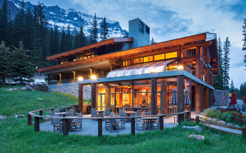 View of Moraine Lake Lodge, a rustic wooden building with large windows, nestled among tall trees with snow-capped mountains in the background. The turquoise waters of Moraine Lake are visible in the foreground, creating a serene and picturesque landscape in Banff National Park, Alberta, Canada.