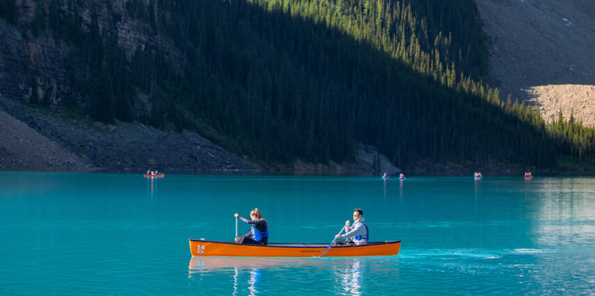 Two people paddle a canoe on the serene, turquoise waters of Moraine Lake, surrounded by towering snow-capped mountains and lush green forests under a clear blue sky.