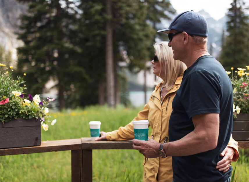 A couple enjoying a coffee together, standing side by side and looking out onto the serene waters of Moraine Lake, surrounded by breathtaking mountain views.
