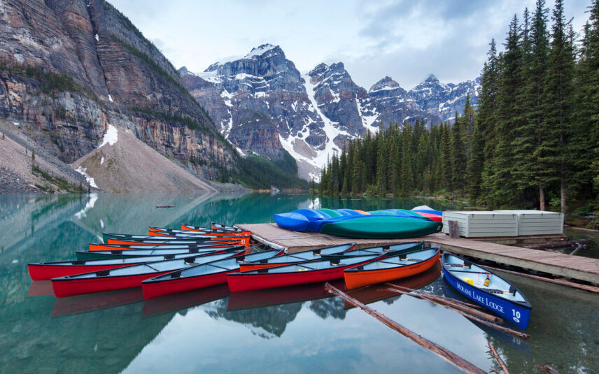 Colorful canoes lined up on the shore of Moraine Lake, with the vibrant turquoise water reflecting the surrounding snow-capped mountain peaks and clear blue sky.
