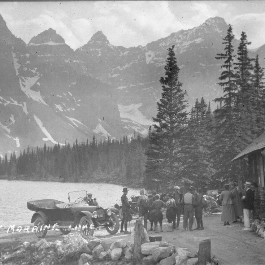 Vintage image of a group of people standing by a car, overlooking the breathtaking view of Moraine Lake and surrounding mountains in Banff National Park.